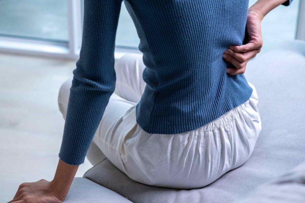 Woman experiencing lower back pain while sitting on a couch, holding her back in discomfort.